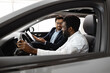 © sofiko14 - Vehicle purchase, customer service, friendly interaction inside car dealership environment. Caucasian car dealer sit inside vehicle with African American buyer, explaining car features.