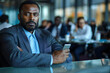 © Vera - African American businessman holding mobile phone looking distracted in a meeting room filled with diverse team employees.