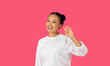 © Prostock-studio - Asian young woman with dark hair tied up in a bun is wearing a white sweater and standing in front of a solid pink background. She is smiling and waving hello with her right hand.