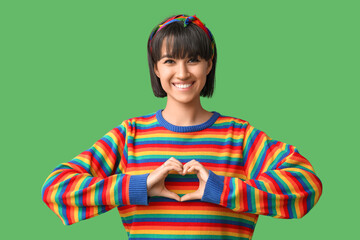 Poster - Beautiful young woman in rainbow t-shirt showing heart gesture on green background. LGBT concept