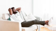 © Prostock-studio - Black man sits on a white couch with his arms behind his head, relaxing and enjoying a moment of peace. His feet are up on a small table beside the couch, and he appears to be taking a break