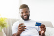 © Prostock-studio - African American man sits on a couch with a bright white background, holding his smartphone in one hand and a credit card in the other, smiling as he makes an online purchase.