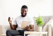 © Prostock-studio - A happy African American man sits on a couch in a living room, holding a tablet in one hand and a mug of coffee in the other. He is smiling and looks relaxed.