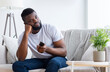 © Prostock-studio - Black man sits on a couch in a living room, looking bored. He is holding a remote control in his hand and has a thoughtful expression on his face. The room is decorated with a houseplant