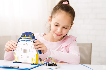 Poster - A young girl in a pink shirt is building a robot toy at home. She is smiling and looks focused on her work. The robot is made of plastic and has blue, white, and yellow parts