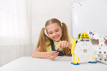 Poster - A young girl, with blonde hair in pigtails, smiles brightly as she connects wires to a robot she is building. She is sitting at a white table in a classroom setting, with a whiteboard behind her.