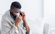 © Prostock-studio - Black man wrapped in a blanket sits on a couch and talks on the phone while blowing his nose. He is clearly sick. He appears to be in his home, as the background is a white wall and a white couch.