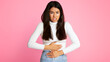 © Prostock-studio - A young Asian woman with long dark hair is standing in front of a pink background. She is holding her stomach with both hands, looking distressed and pained.