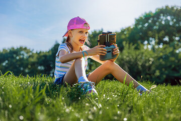 Girl 8-9 years old is operating the drone by remote control in the park. Kid sitting in grass, hold in hands controller, playing with quadcopter with angry face expression