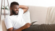 © Prostock-studio - African American man with a beard is sitting on a couch, leaning back, while using a tablet. He is wearing a white shirt and gray pants. He is looking to the side with a smile on his face.