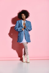 A fashionable African American woman with curly hair standing confidently in front of a vibrant pink wall.