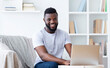 © Prostock-studio - Black man with a beard sits on a couch in a home office, using a laptop computer. He is smiling and looks happy and relaxed. Room is decorated with white walls and a bookcase full of books.