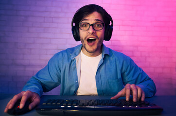 Leinwandbilder - A young man wearing headphones and glasses is seated in front of a keyboard, looking surprised with his mouth open. A brick wall is behind him, and the room is lit with purple and pink lighting.