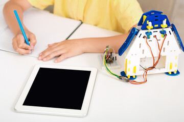 Poster - A child is sitting at a desk, learning about robotics and coding. They are using a tablet computer and a robot kit. The childs hand is shown writing on a piece of paper