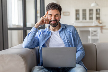 Poster - A man is sitting on a light gray couch while using a silver laptop. He is wearing a light blue button-down shirt over a white t-shirt. He is smiling and appears to be happy.