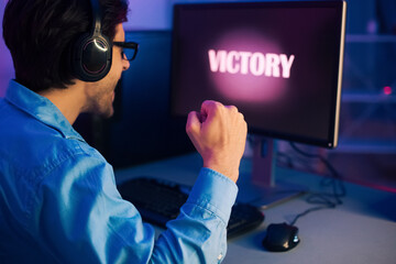 Aufkleber - A young man wearing headphones sits in front of a computer screen, his fist clenched in the air, celebrating a victory in a video game. The screen displays the word Victory in a bright pink glow