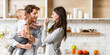 © Prostock-studio - A family of three enjoys a meal together in a modern kitchen. The father holds their young child, while the mother feeds them with a spoon. They are all smiling and seem to be enjoying their meal.