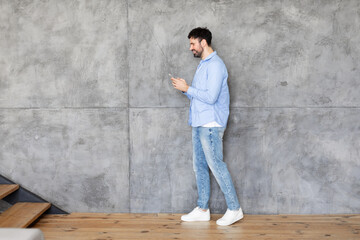 Poster - A man in a blue shirt and jeans is standing near a concrete wall with wooden stairs. He is looking at his smartphone and smiling. The room appears to be modern and spacious.