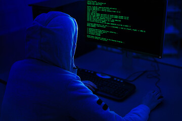Fototapete - A person wearing a hooded sweatshirt sits in front of a computer with a dark background. Computer screen displays green code, indicating that the person is working on a programming or technical task.