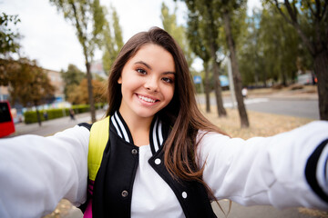 Wall Mural - Portrait of adorable optimistic girl with brunette hairdo wear uniform rucksack making selfie smiling on green city street outdoors