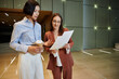 © Bliss - Two businesswomen stand in a modern office lobby, discussing paperwork while holding coffee cups.
