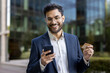 © Liubomir - Happy businessman standing outdoors holding smartphone and credit card, making mobile payment. Professional wearing formal suit smiling while using technology for online transaction.