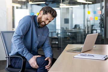 Wall Mural - A young man sits in the office at the desk and holds his hands on his knee, grimaced and feels severe pain in his leg