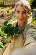 © pressmaster - Happy female farmer with green strawberry seedling in small pot looking at camera during livestream and communicating with online audience