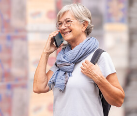 Sticker - Smiling senior white haired Caucasian woman tourist using mobile phone while walking in Seville, Spain holding a backpack enjoying vacation trip freedom, healthy lifestyle in retirement