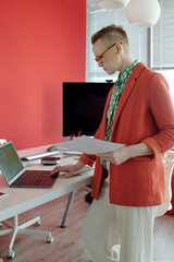 Businesswoman holding documents and using laptop in modern office with red walls while working in advertising agency