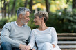 © Sunday Cat Studio - Happy elderly couple sitting on park bench, looking at each other lovingly, surrounded by lush greenery, enjoying peaceful moment together.