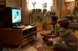 © pressmaster - Two multiethnic boys engaged in playing a retro video game on an old television set, sitting on a carpeted floor with plants and home decor in the background