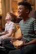 © pressmaster - Vertical shot of two multiethnic schoolboys laughing and watching TV while sitting on floor in cozy living room, enjoying time together with popcorn and a relaxed atmosphere