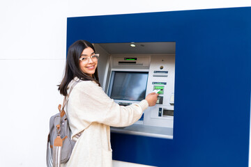 A cheerful woman with a backpack uses an ATM, smiling while inserting her card. emphasizes convenience and positive financial interactions. Finance and technology concept.