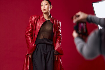 Wall Mural - Fashion asian woman in red leather coat and black pants taking a photo in front of red background