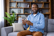 © Liubomir - Confident man sitting on sofa holding digital tablet in living room. Relaxing and enjoying leisure time. Background includes bookshelves and green plants. Comfortable and cozy home environment.
