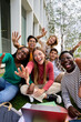 © CarlosBarquero - Vertical. Amusing portrait of group diverse young friends excited waving at camera with hands sitting on campus grass. Gen z of happy students gathered posing together for photo outside faculty class