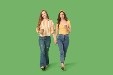 Wall Mural - Beautiful young sisters with cups of coffee walking on green background