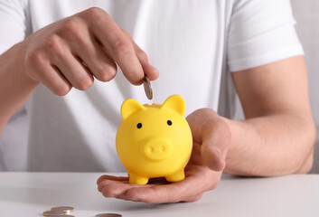 Poster - Man putting coin into yellow piggy bank at white table, closeup