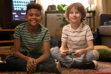 Portrait of two diverse boys smiling, looking at camera and sitting on carpeted floor while playing retro video games during free time at home