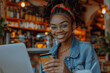 © btiger - Happy young black woman shopping online with credit card and smartphone in coffee shop, wearing glasses and denim jacket