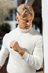 A handsome blonde man poses on a street in Florida wearing a white sweater.