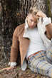 © Bliss - A handsome blonde man wearing a brown suede jacket and plaid pants in park near tree