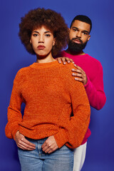 A young African American man and woman, friends, stand together in vibrant casual attire against a blue background.