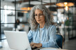 © btiger - Mature female entrepreneur working on laptop in modern office, older businesswoman with grey curly hair and glasses typing on computer at desk. Professional woman managing digital project