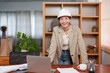 © Ekkasit A Siam - Asian female designer standing at a wooden desk, smiling and leaning forward. Wearing a white hard hat and a beige blazer in a modern office setting. Laptop, coffee cup, and documents on the desk.