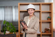 © Ekkasit A Siam - Asian female designer holding a tablet, smiling and wearing a white hard hat. Standing in a modern office setting with wooden shelves in the background. Casual attire with a beige blazer.