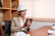© Ekkasit A Siam - Asian female designer seated at a wooden desk, holding a tablet and smiling. Wearing a white hard hat and a beige blazer in a modern office setting. Documents and coffee cup visible on the desk.