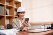 © Ekkasit A Siam - Asian female architect wearing a white hard hat and a beige blazer, smiling while using a smartphone at a wooden desk. Modern office setting with documents, coffee cup, and tablet visible.