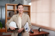 © Ekkasit A Siam - Asian female architect smiling while holding a white hard hat and rolled-up plans. Wearing a beige blazer in a modern office setting with wooden shelves. Positive and professional expression.